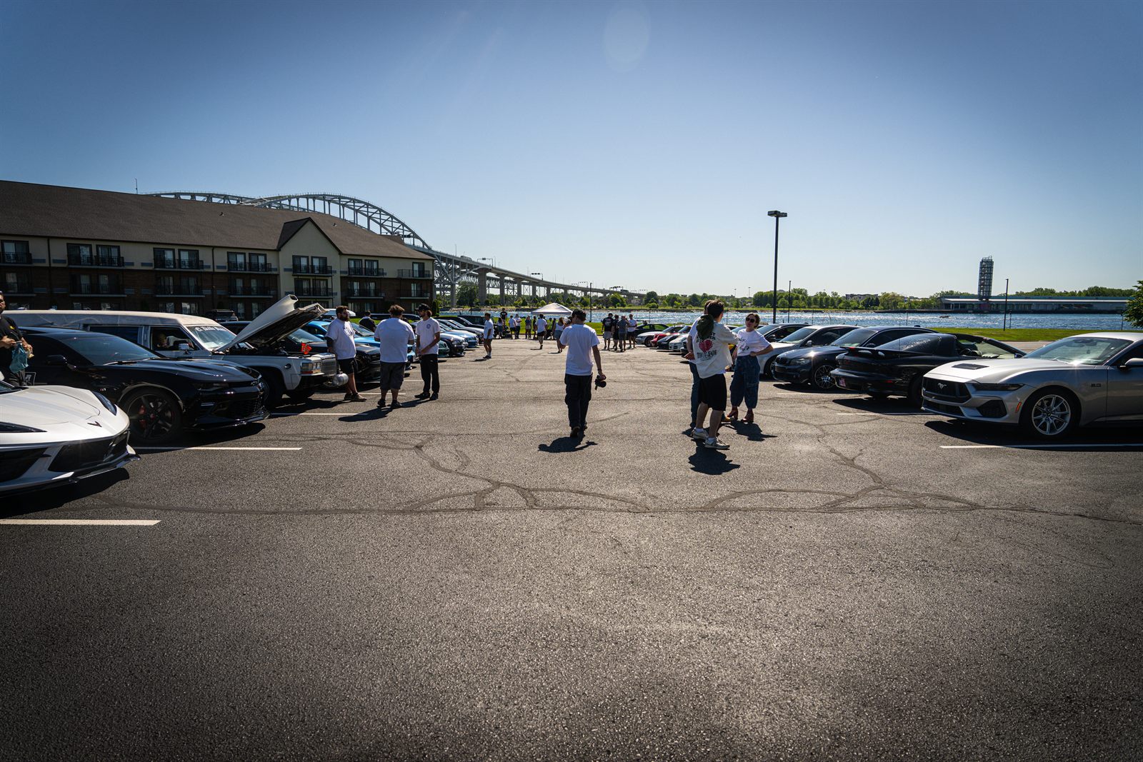 A Baseline Rally meet on a waterfront parking lot, with rows of cars, attendees walking between them, and the Blue Water Bridge in the background.