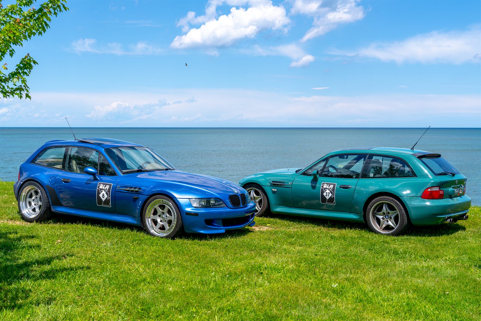 Two BMW Z3 M Coupes, one blue and one teal, parked on the grass overlooking Lake Michigan, both wearing Baseline Rally door decals.