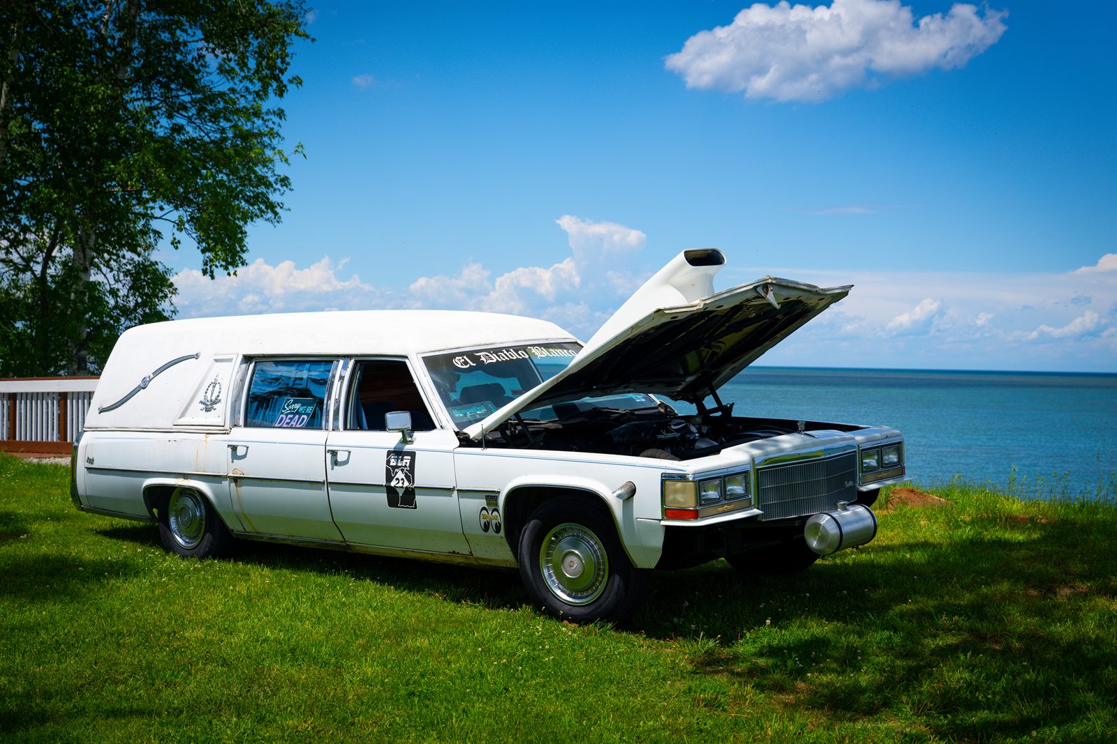 A white Cadillac hearse lettered "El Diablo Blanco" with its hood raised, wearing a Baseline Rally door decal, parked on the grass overlooking the lake.